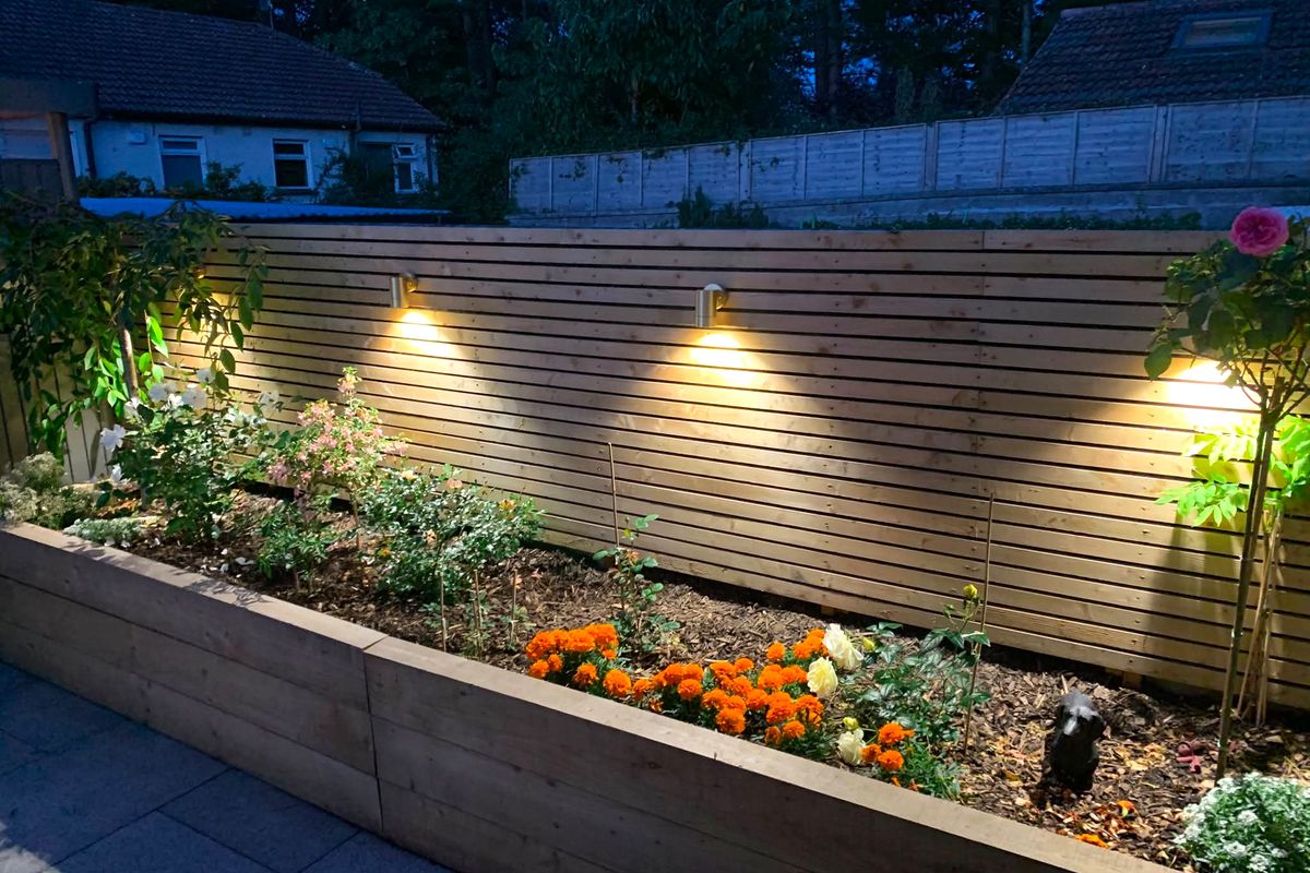 Warm downlights along a slatted timber fence above a planted raised bed at dusk