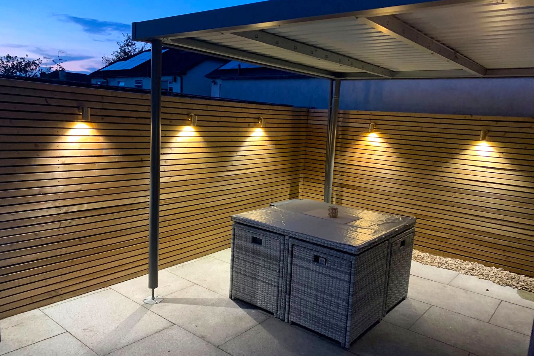 A Dublin patio at dusk, five warm downlights along a slatted timber fence under a canopy