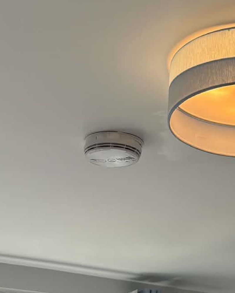A smoke alarm fitted flush to a bedroom ceiling, beside a lit drum pendant