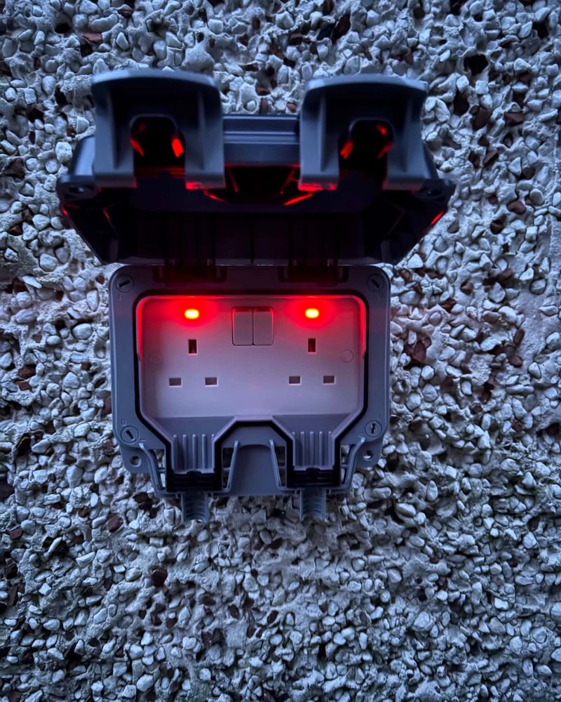A weatherproof outdoor double socket with red indicator lights, at dusk